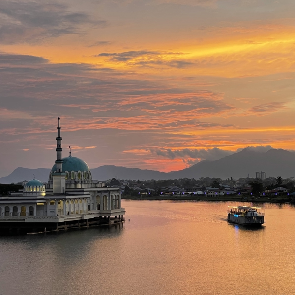 Smuk solnedgang over floden i Kuching med en moské ved vandkanten, en turistbåd på floden og bjerge i baggrunden under en orangegylden himmel